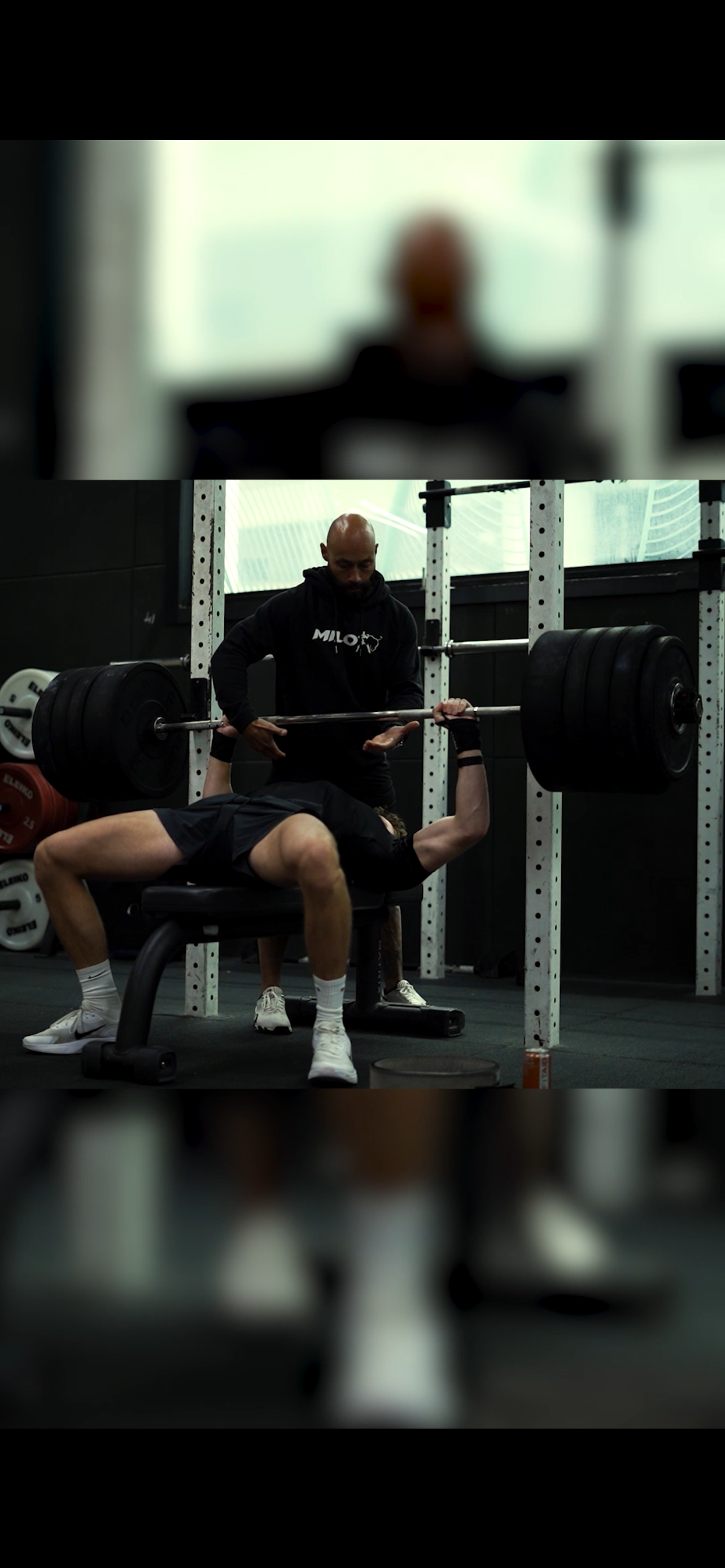 Een man ligt op een bankje terwijl hij een grote halter met gewichten omhoog duwt onder toezicht van een trainer in een fitnessruimte.