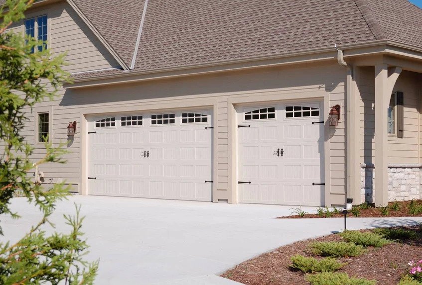 A beige house with a three-car garage with white doors and black hardware, a landscaped front yard, and a concrete driveway.
