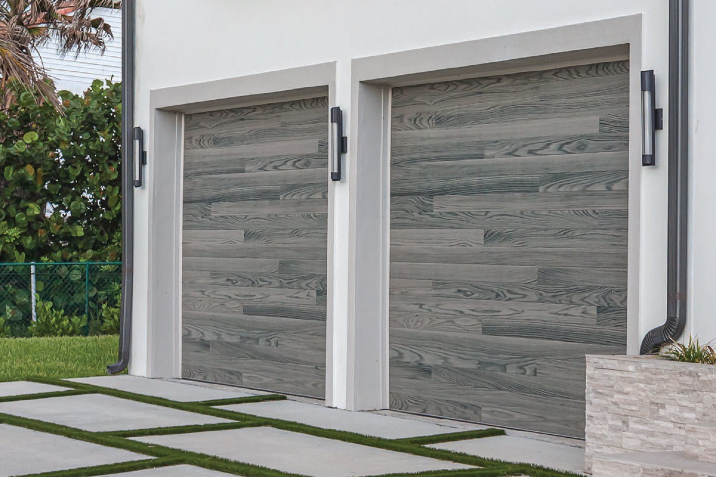 Double garage doors with a wood grain finish, in a modern home exterior, with concrete stepping stones and a landscaped yard.