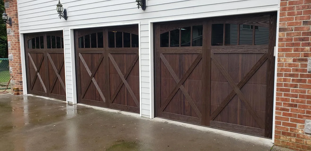 Three brown sectional garage doors with windows at the top, installed on a white house with brick accents. Two outdoor wall lanterns are mounted on the house.