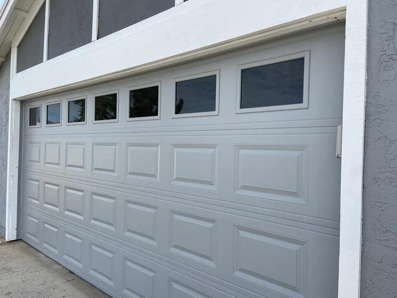 White sectional garage door with rectangular windows at the top, installed on a gray house with a concrete driveway.