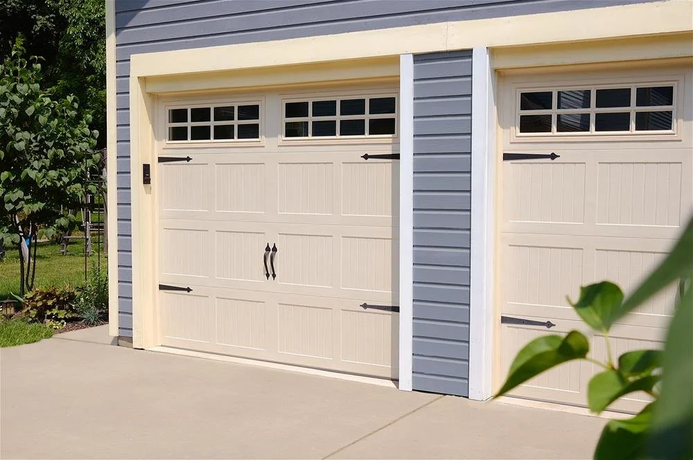 Two beige garage doors with black hardware in a blue house with white trim, surrounded by greenery.
