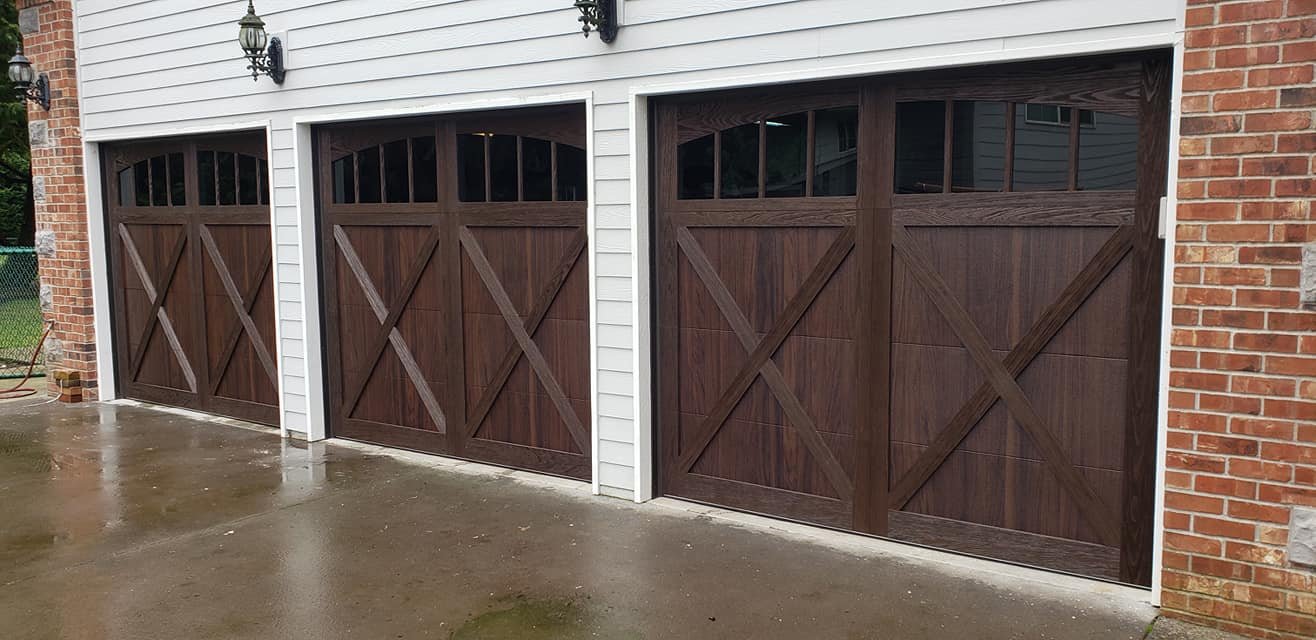 Three brown garage doors with crossbuck design, set in a white and brick building, with black outdoor lanterns mounted above each door.