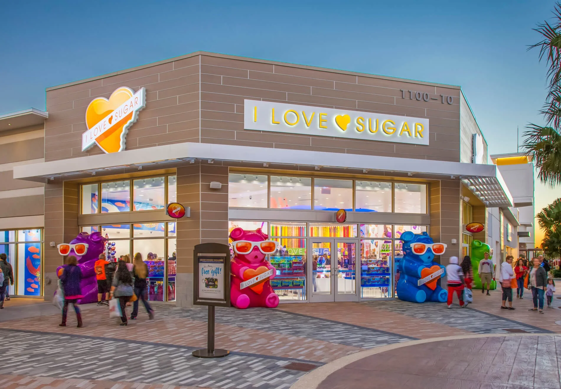 Exterior of a store named 'I Love Sugar' with large bear statues wearing sunglasses in front, colorful shopping bags, and people walking outside during sunset.
