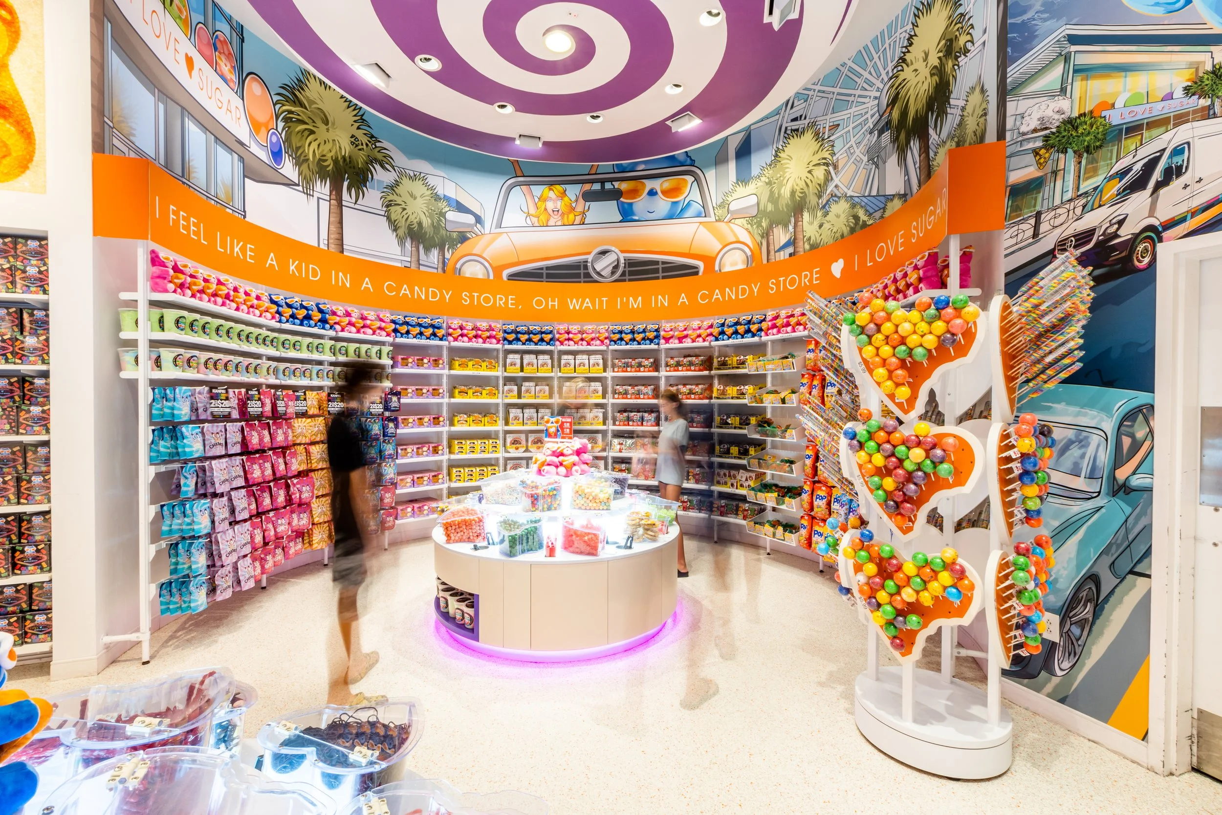 Our Myrtle Beach store with colorful shelves filled with various candie, decorated with a mural of a cartoon convertible with our Gummy Bear driving, palm trees, and Skywheel in the background.