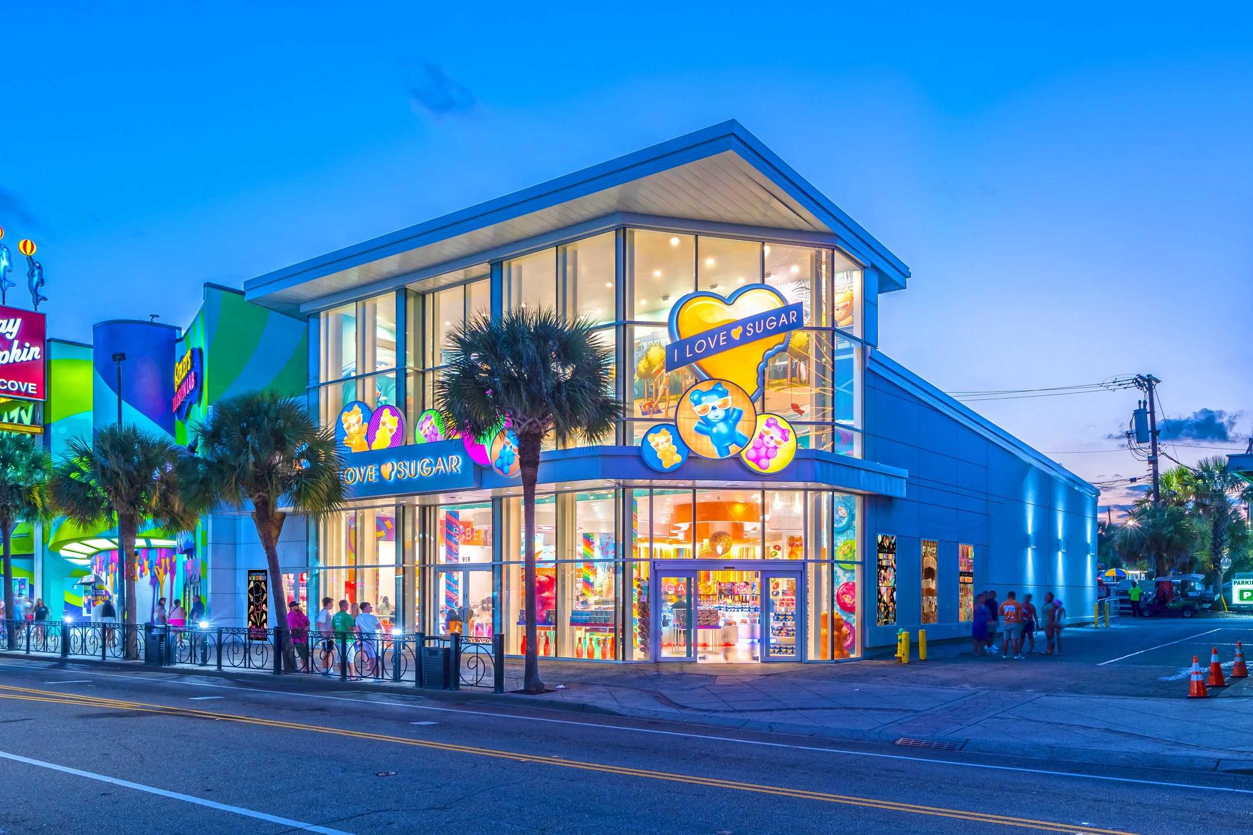 Our brightly lit, modern two-story Myrtle Beach I LOVE SUGAR storefront with colorful signage and large glass windows, palm trees in front, and people walking nearby during evening hours.