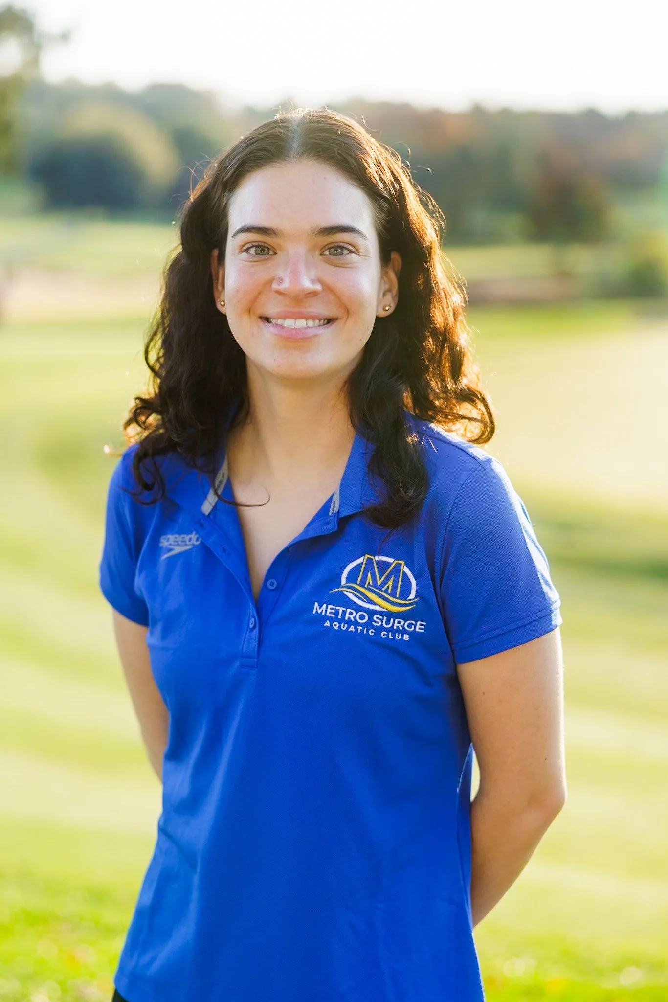 A young woman in a blue polo shirt standing outdoors near a body of water, with trees and greenery in the background, smiling at the camera.