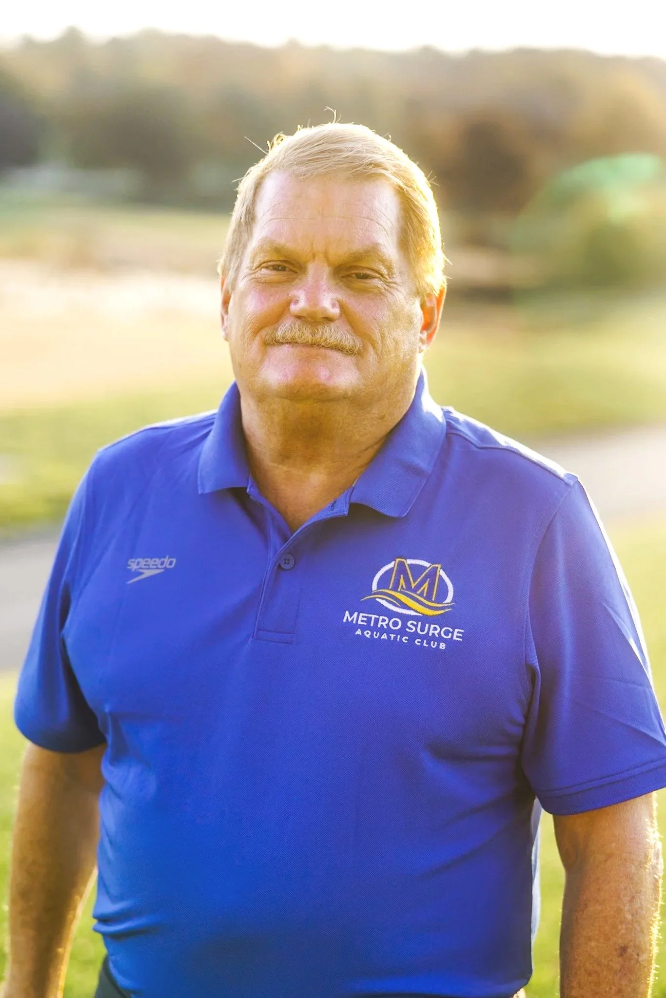 A middle-aged man with light hair, mustache, and tan skin, wearing a blue polo shirt with 'Metro Surge Aquatic Club' and the Speedo logo, standing outdoors with blurred trees and grass in the background, sunlight shining behind him.