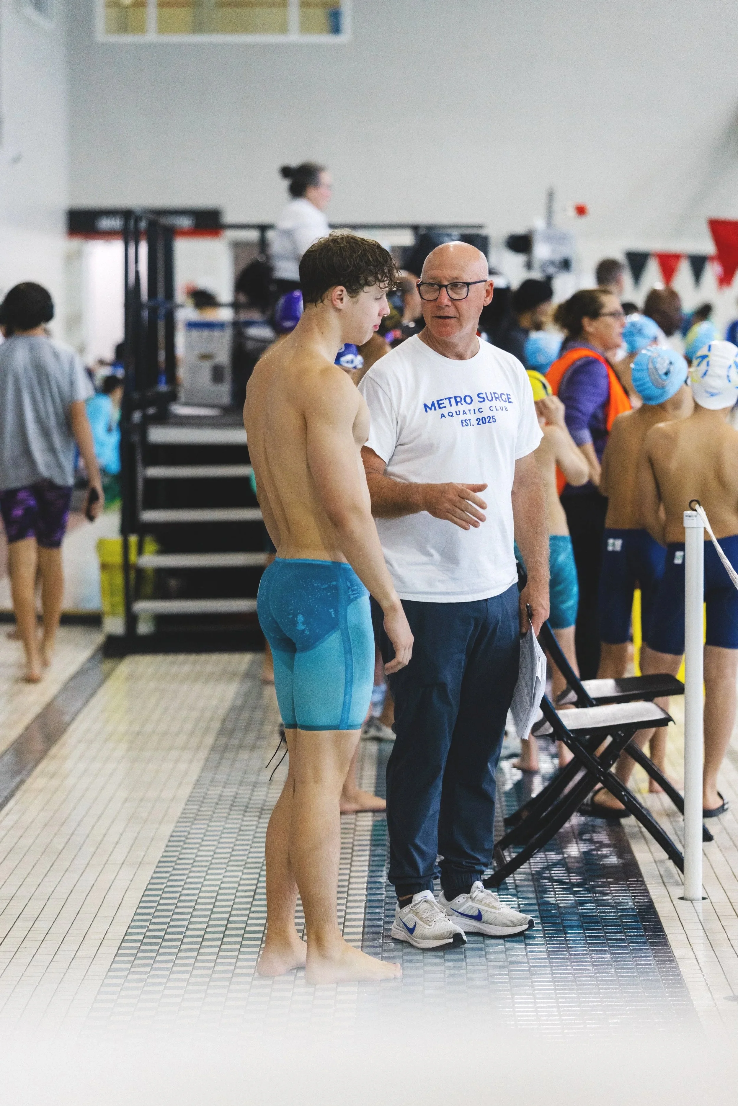 A young male swimmer in blue swim shorts talking to an older man in a white T-shirt and glasses at an indoor swim meet.
