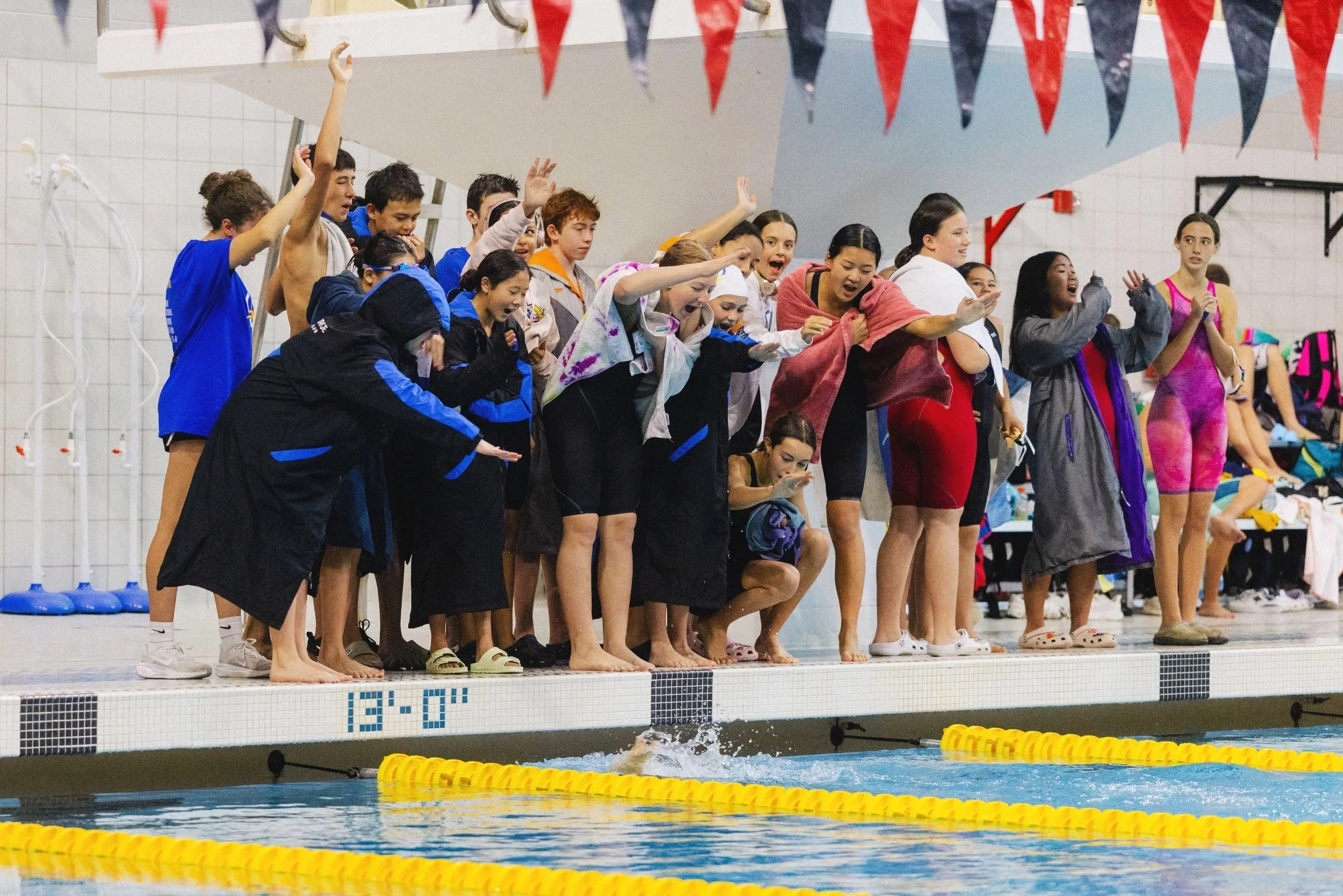 Swimmers and spectators at an indoor swimming competition, with some children on the pool deck cheering and some preparing to swim.