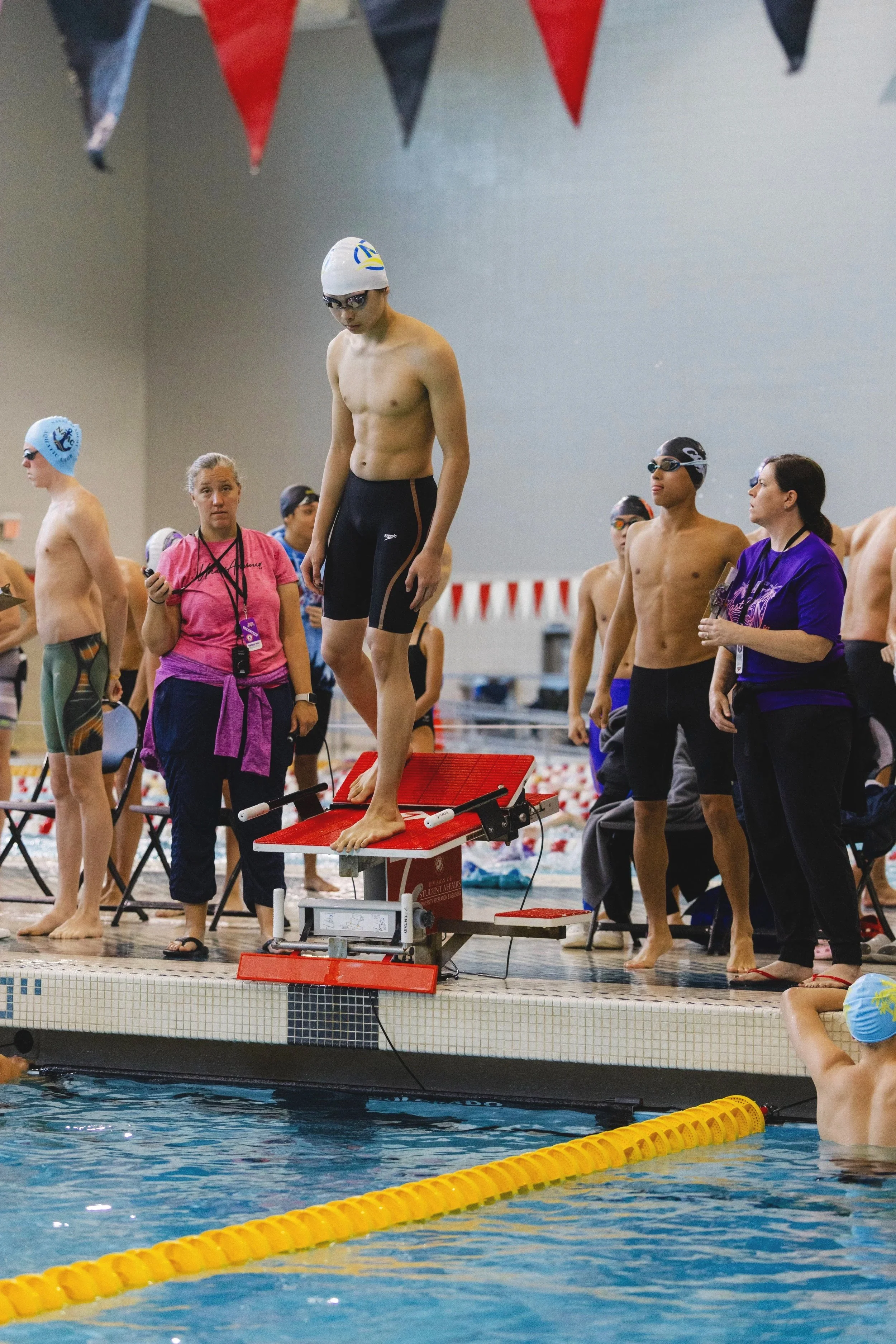 Swimmers on starting blocks at an indoor swimming competition, with coaches and officials watching.