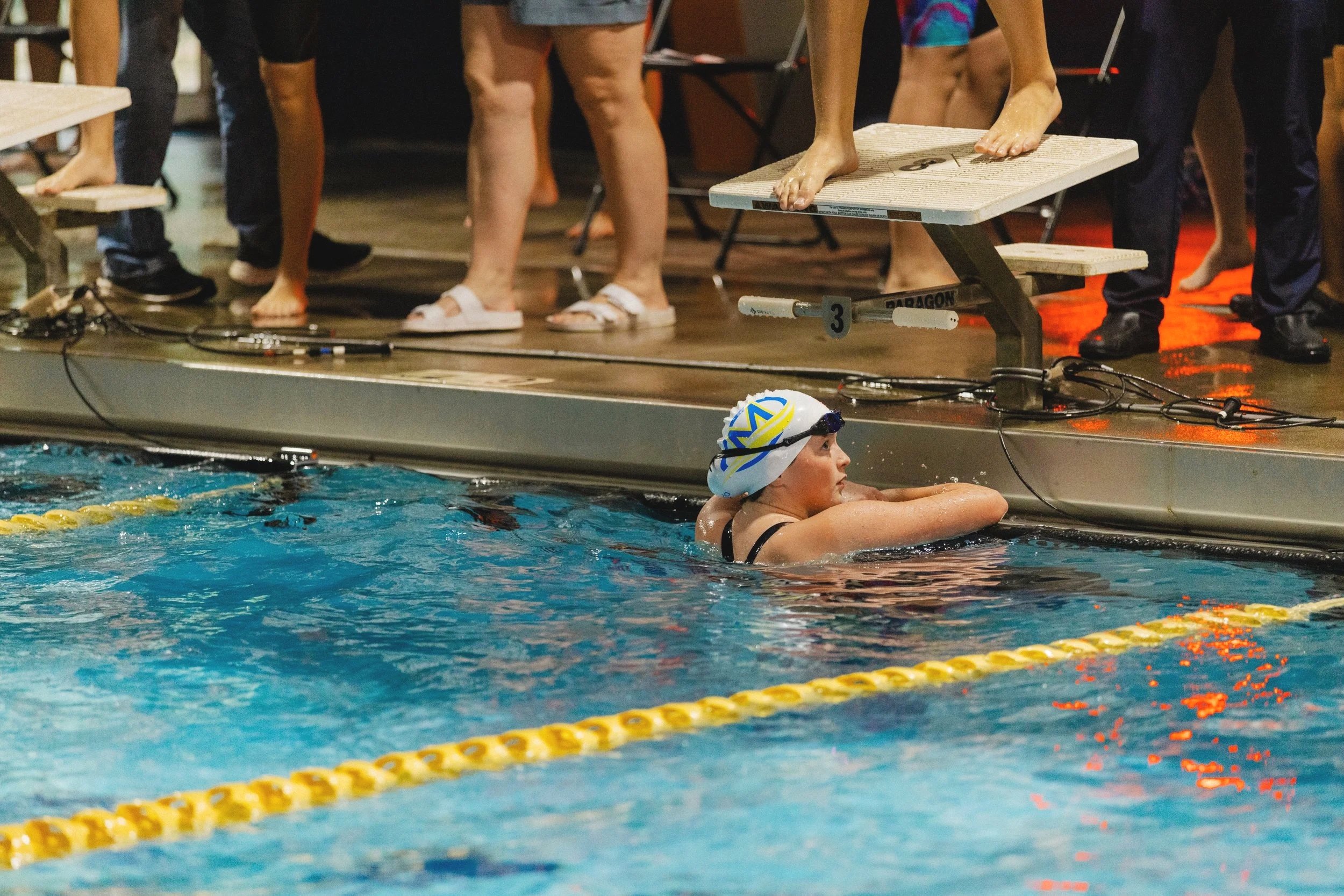 Swimmer resting with arms on the edge of the pool, wearing a swim cap and goggles, next to the starting block at a swimming competition.