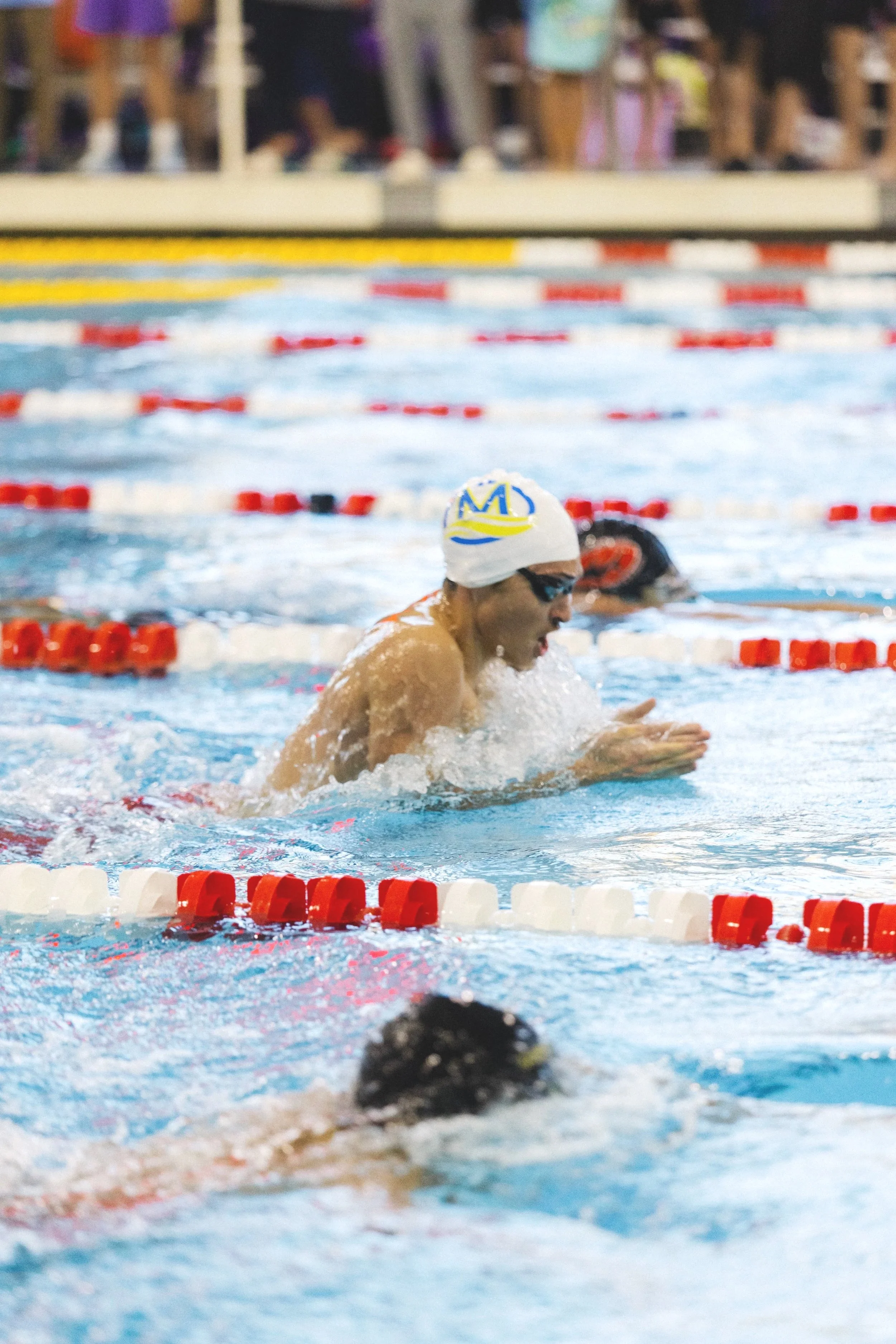A swimmer wearing a white swim cap with a yellow and blue crown emblem, black goggles, and a swimsuit, emerges from the pool with water splashing around. The swimmer is in a lane separated by red, white, and yellow lane dividers. In the background, another swimmer is partially visible, along with blurred spectators or officials behind a glass or barrier.