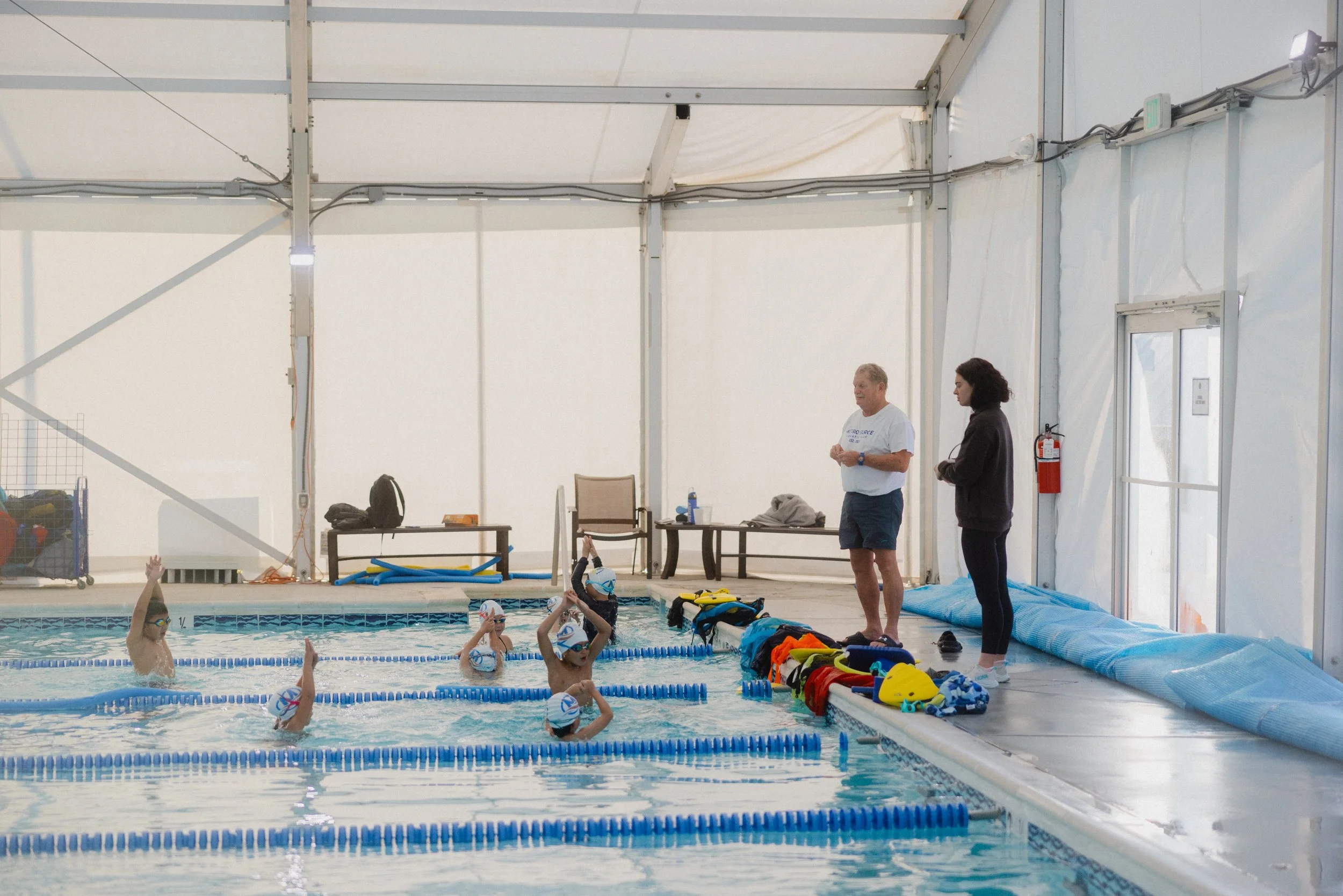 Swimmers in an indoor pool during a swim lesson with two instructors standing by the poolside, various swimming gear placed on the edge.