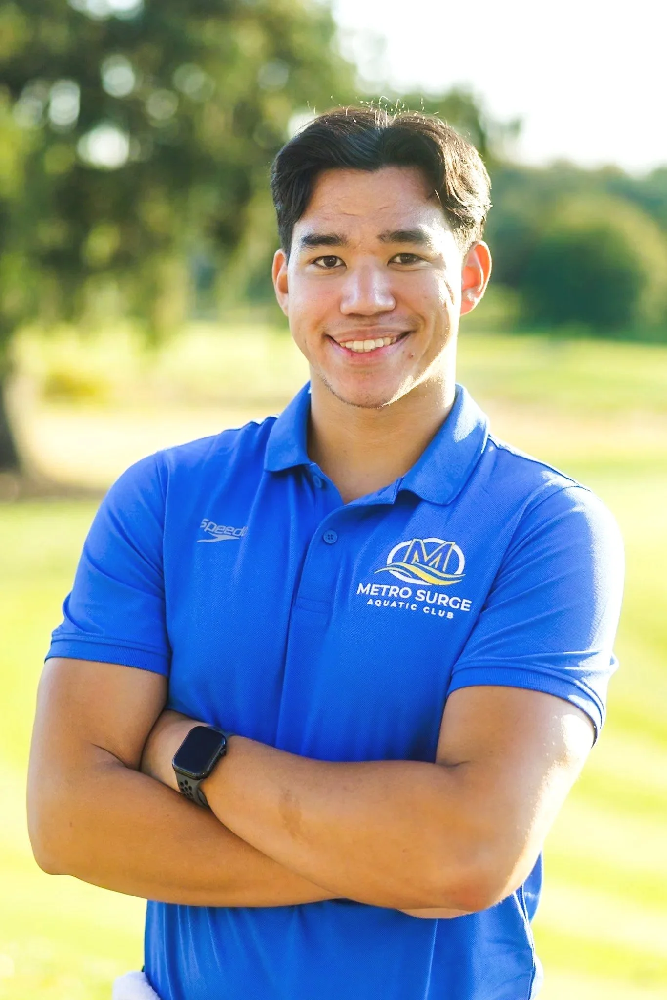 Young man with crossed arms smiling outdoors, wearing a blue polo shirt with 'Metro Surge Aquatic Club' logo, and a black smartwatch, with blurred trees in the background.
