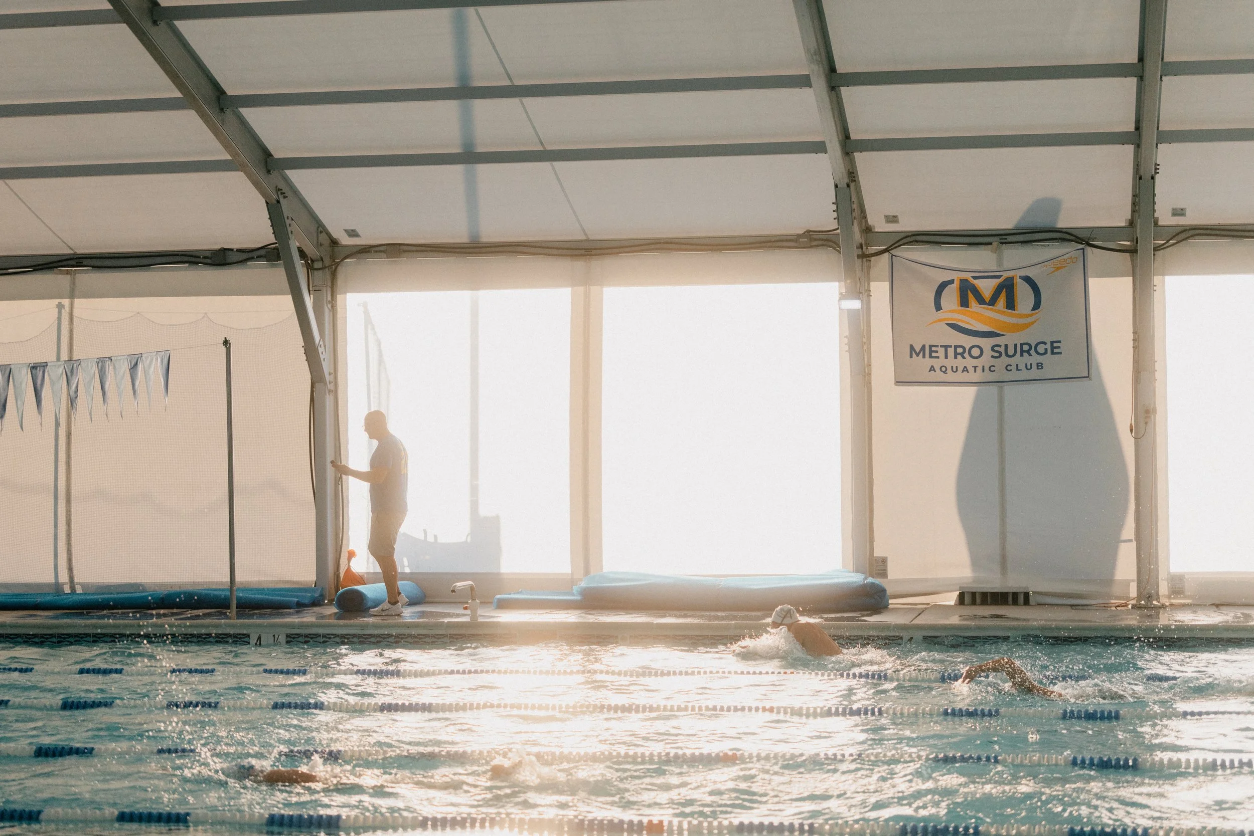 Indoor swimming pool at Metro Surge Aquatic Club with swimmers practicing and a lifeguard standing by the poolside.