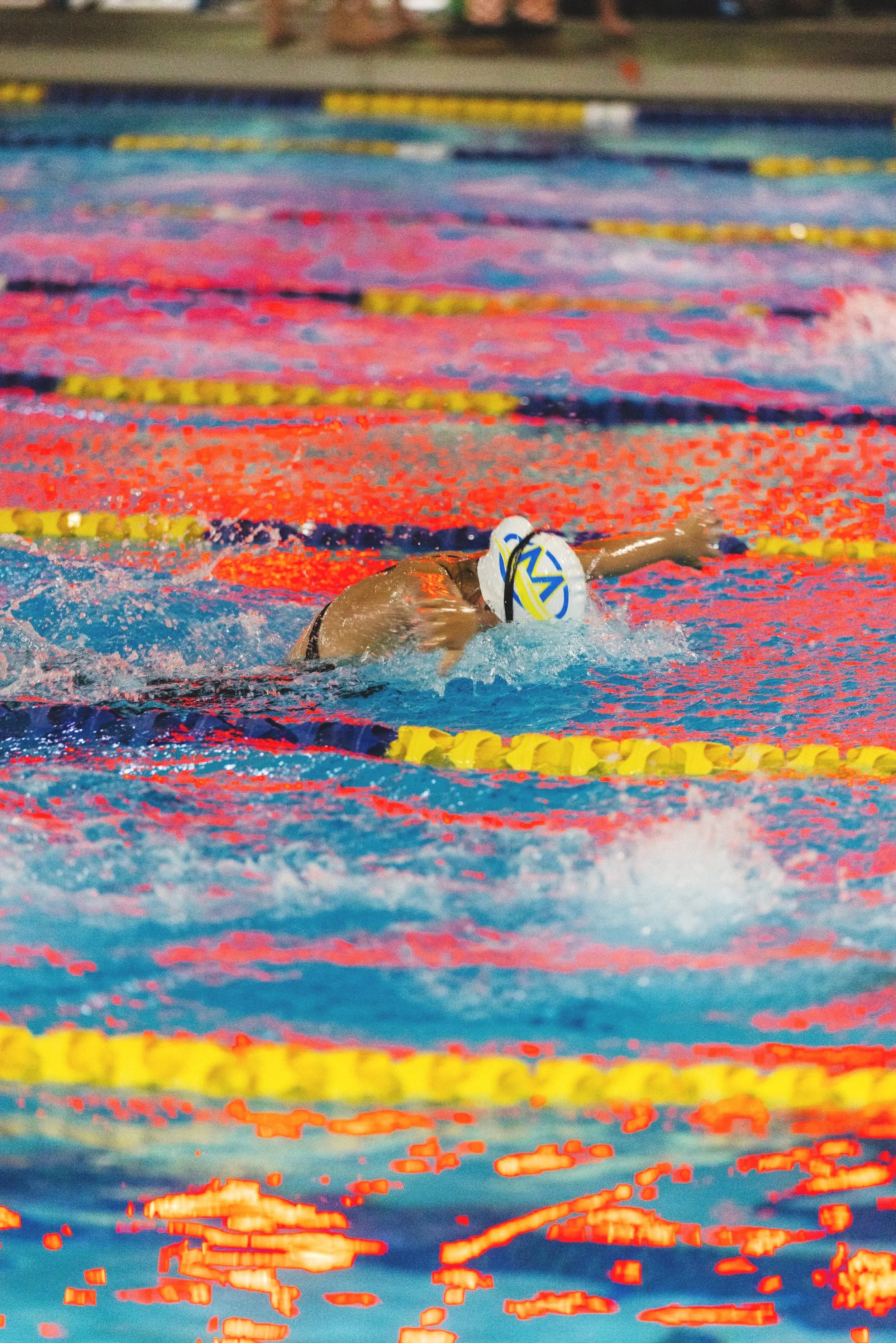 Swimmer in a pool competing in a race, wearing a white swim cap with yellow and blue markings, moving through the water with arms extended.