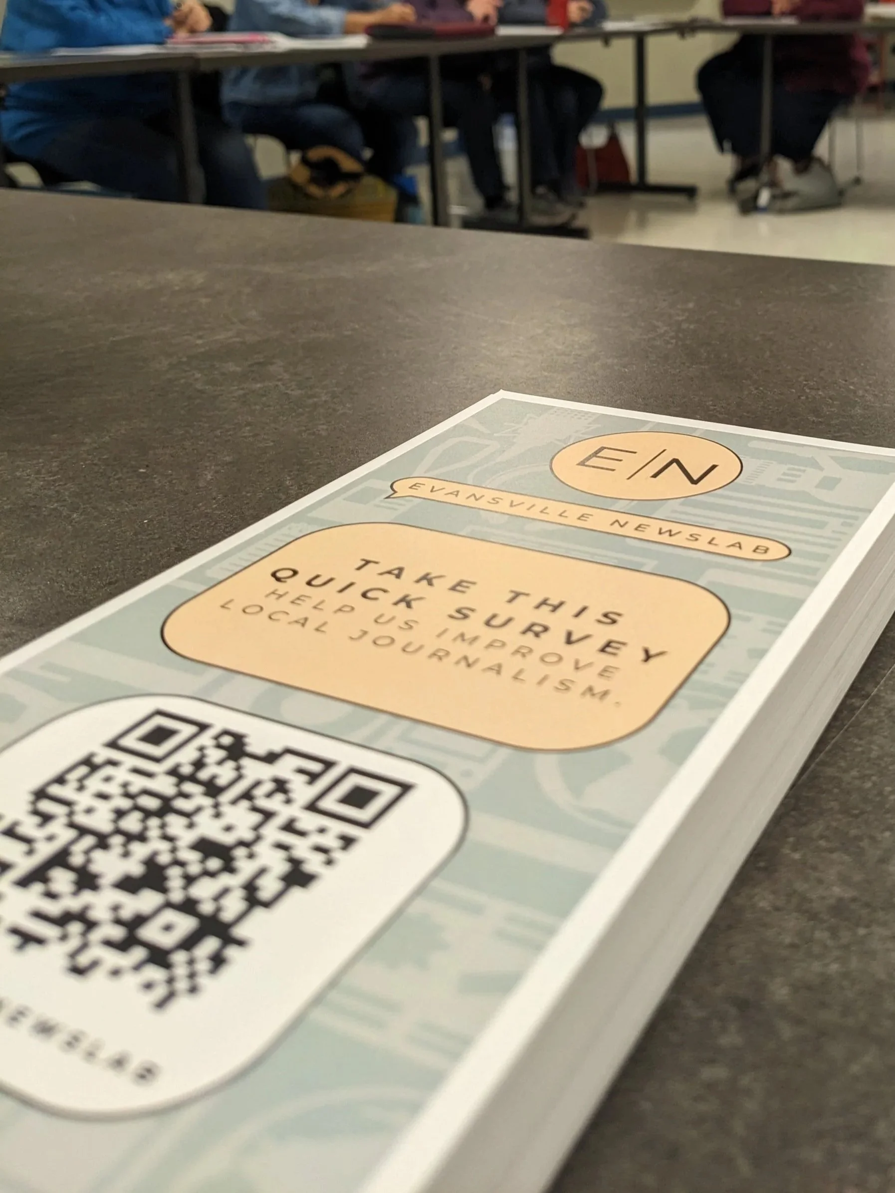 A pamphlet on a table with the headline 'EIN' and instructions to take a quick survey to improve local journalism, featuring a QR code at the bottom. The background shows a classroom with students sitting at desks.
