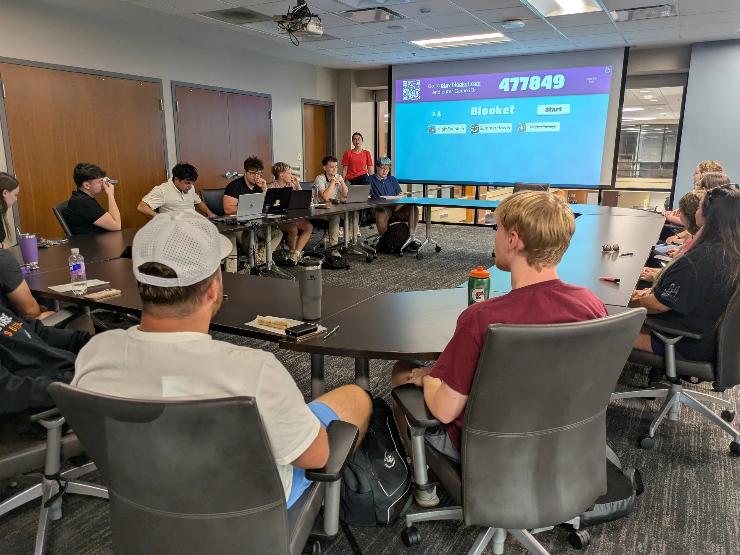 A group of young people sitting around a large, curved conference table in a modern room with a large screen displaying a game scores and instructions. Some are using laptops, while others listen attentively.