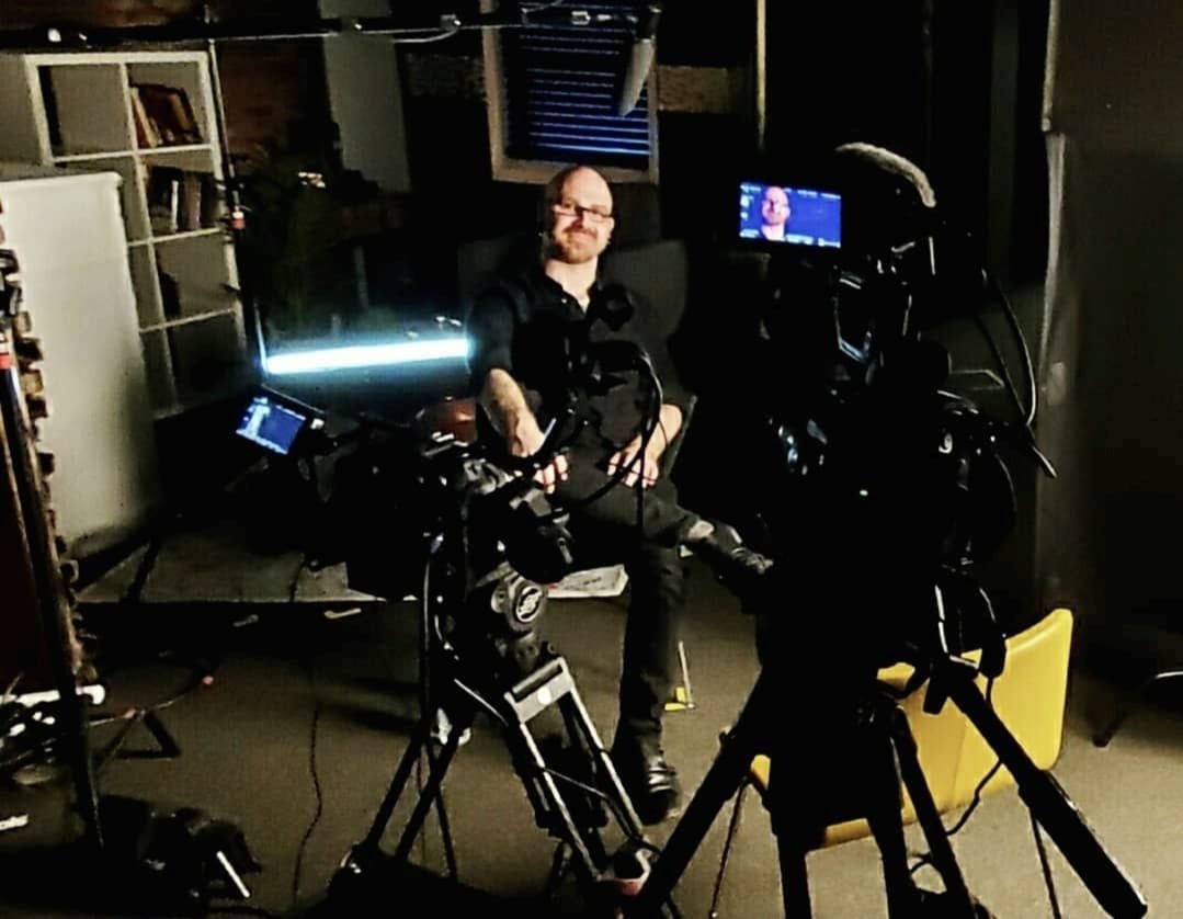 Shea Bilé sits in a dimly lit room surrounded by professional video production equipment, including cameras, monitors, and lighting. Shea is smiling and looking at the camera.