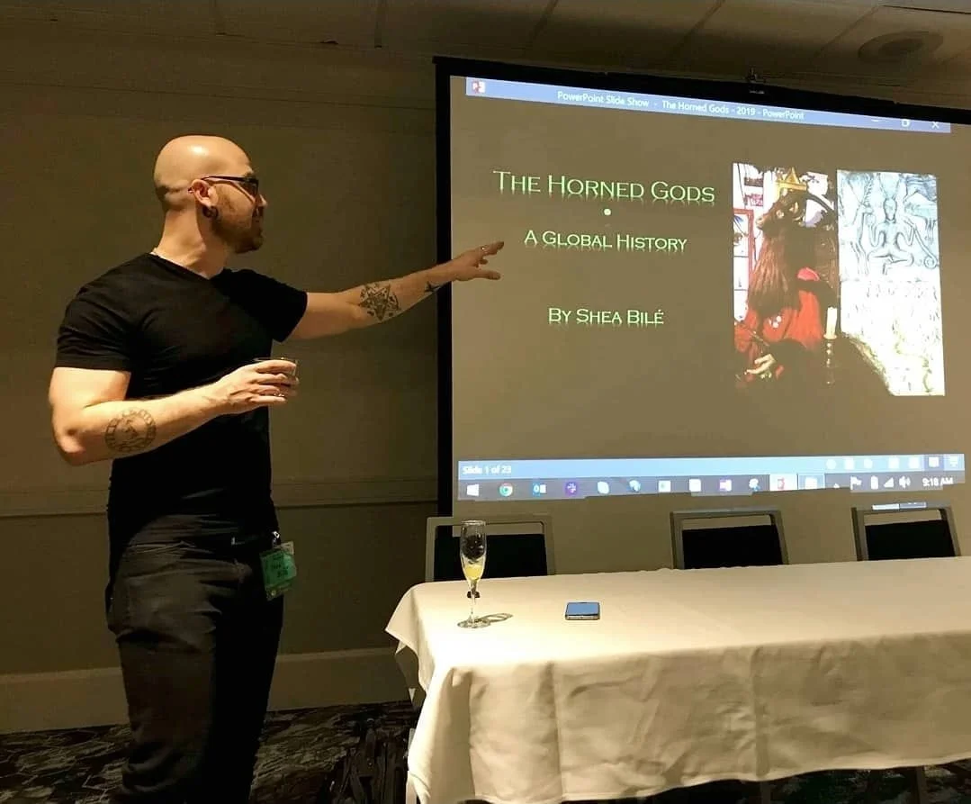 Shea Bilé wears a black t-shirt and is pointing at a presentation slide projected on a screen, with a glass of champagne on the table in front of him. Pantheacon. 
