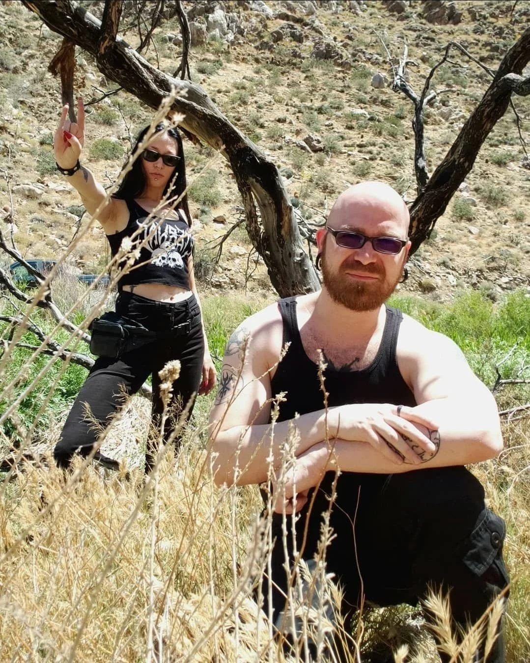 Shea Bilé and Luzia Lowe are outdoors in a dry, rocky terrain with sparse vegetation and a leafless tree. They are wearing dark clothing, sunglasses, and have tattoos. Luzia is standing showing sign of horns while Shea is squatting.