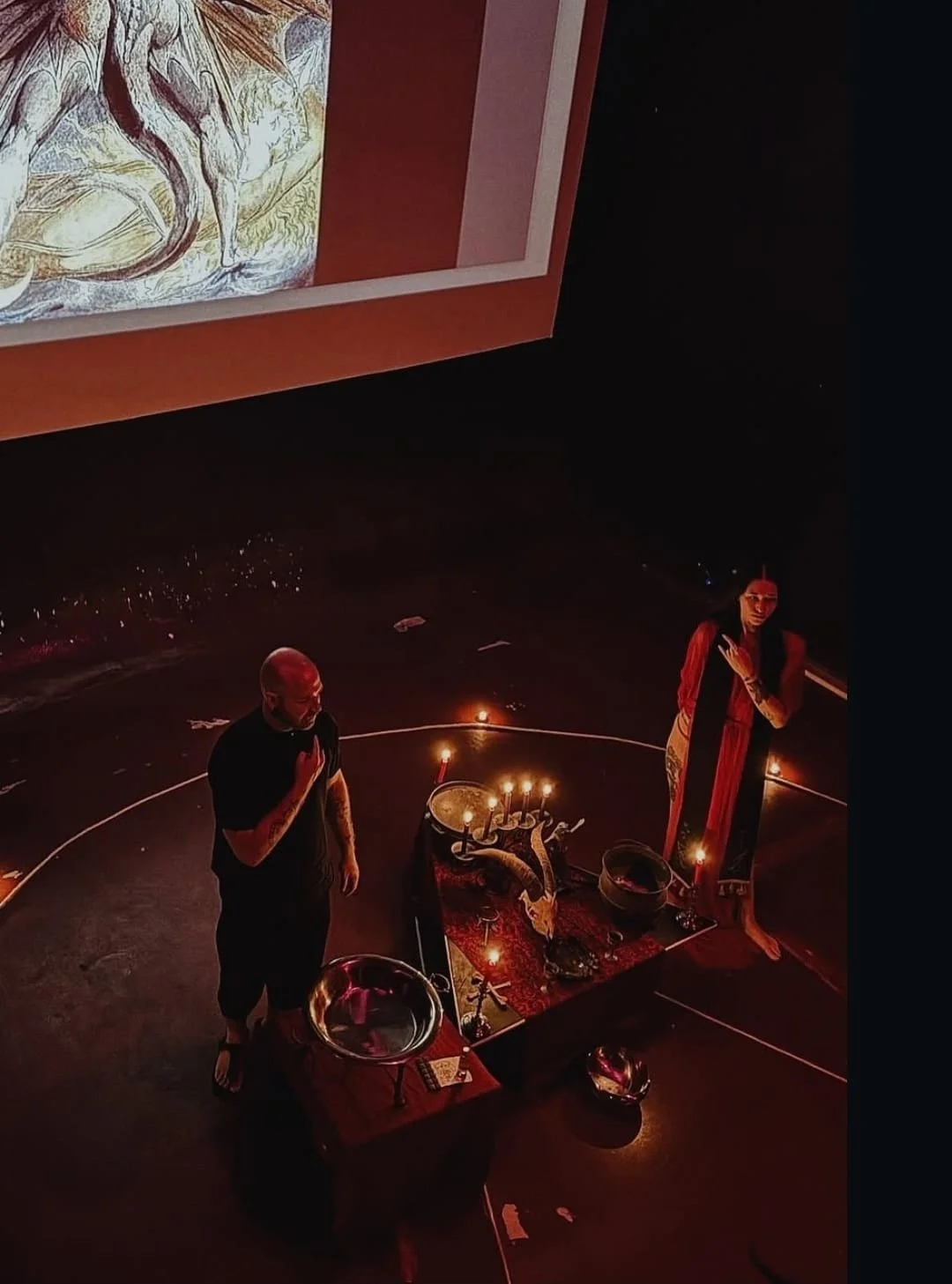 Shea Bilé and Luzia Lowe perform a ritual, standing on a dark floor around a table with candles, bowls, and decorative objects, with a large image of The Great Red Dragon by William blake projected on the wall behind them.
