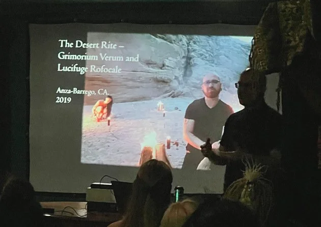 Shea Bilé conducts a lecture in a dark room. A presentation slide behind him depicts a picture of him and Luzia Lowe conducting a ritual in a desert.
