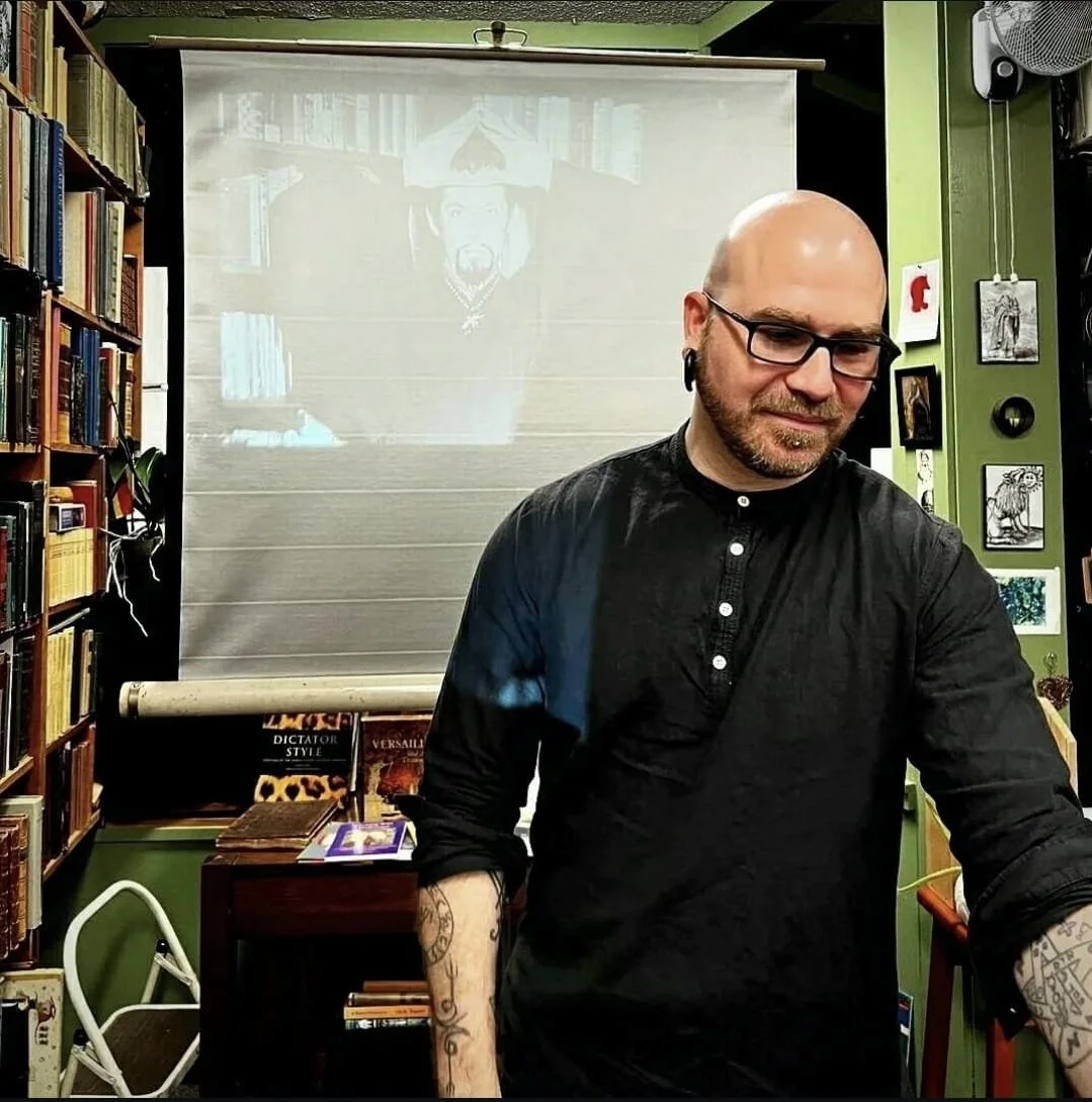 Shea Bilé stands in front of a bookshelf, with a projector screen behind him displaying an image of Anton LaVey. Splendor Solis. 