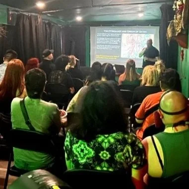 A group of people attending a lecture by Shea Bilé in a dark room at the Crooked Path in Los Angeles, CA, and a large screen displaying a presentation slide.