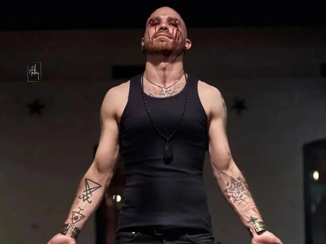 Shea Bilé stands with a serious expression, wearing a black tank top and various necklaces, with blood on his arms and face.