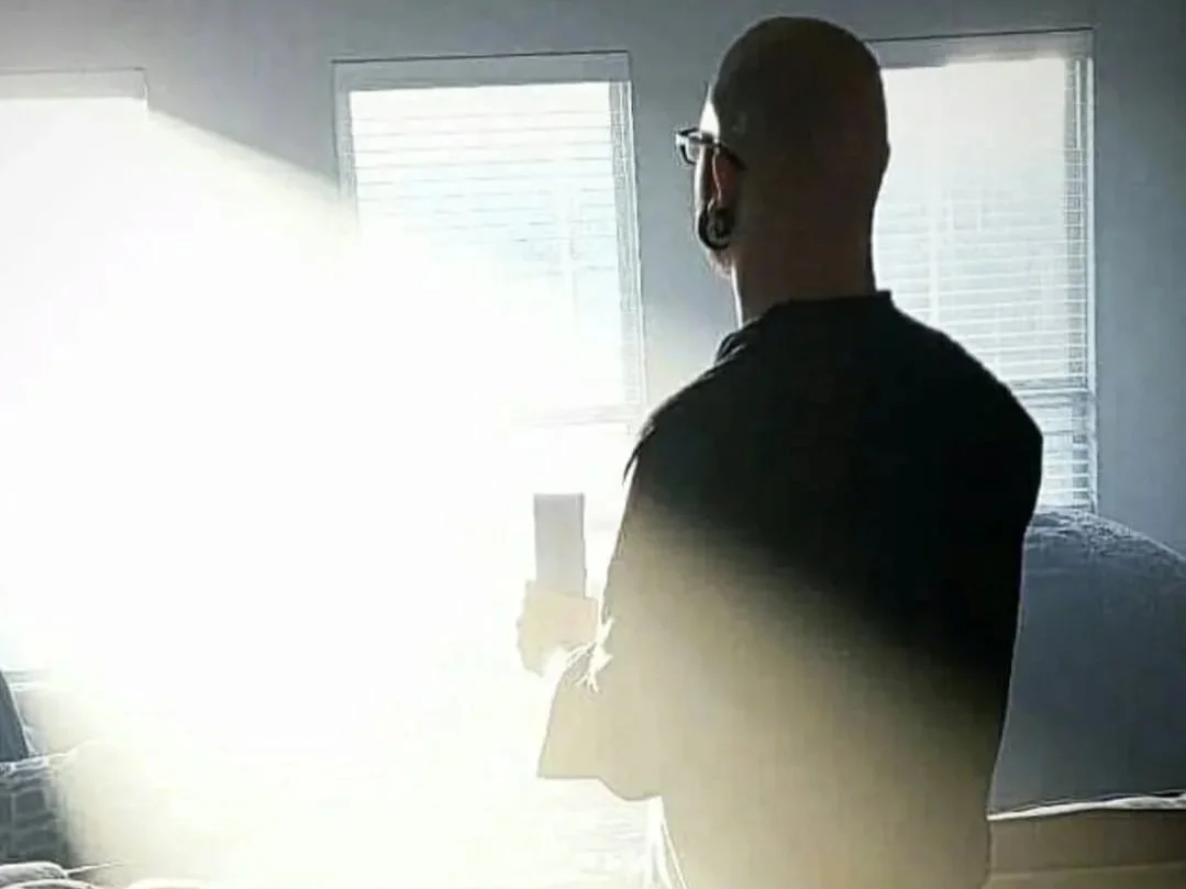 Shea Bilé is standing in front of a few brightly lit windows, one beaming a bright ray of light. He holds a candle. 