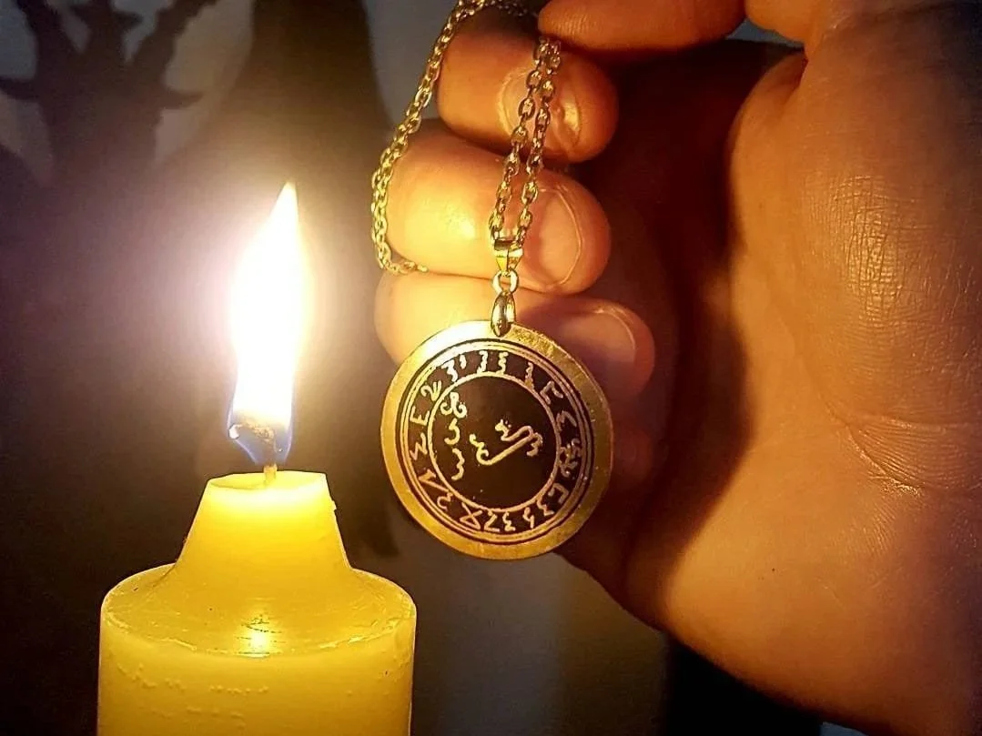 Close-up of a hand holding a gold chain with a circular pendant featuring a circular sigil of Lucifer. A candle with a yellow wax and a flame is visible in the background.