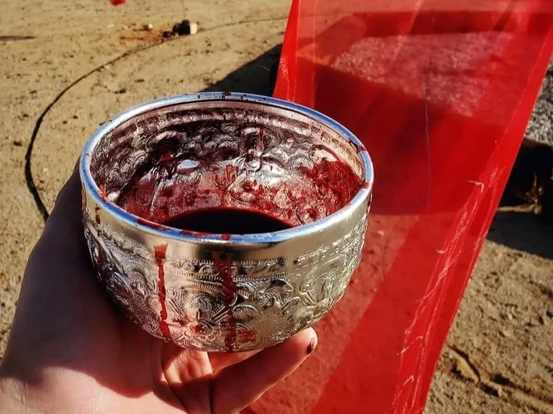 A hand holding a decorative metal bowl with blood, set against a sandy outdoor background with a visible red banner.