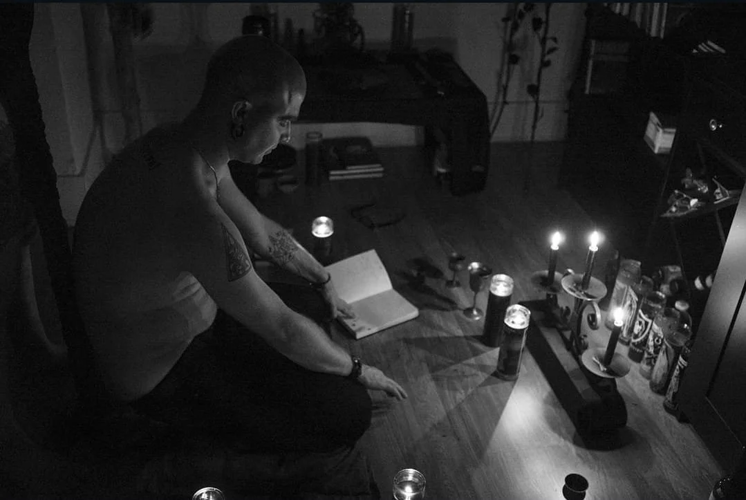 Shea Bilé sits on the floor with lit candles and books in a dimly lit room.