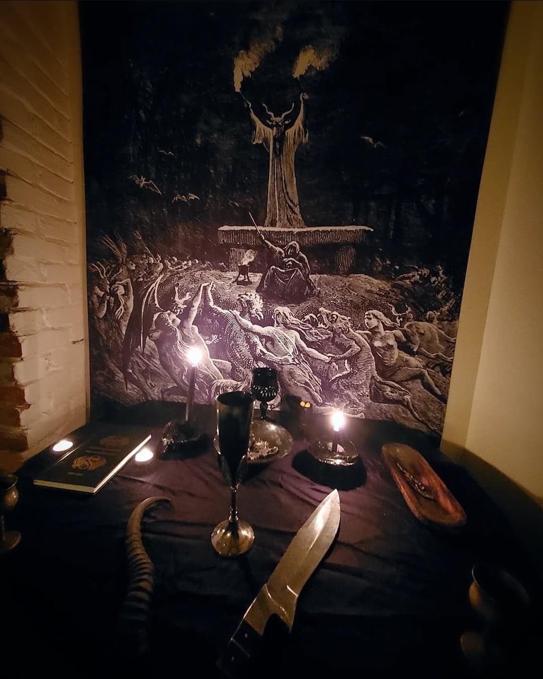 A dark scene resembling a satanic ritual setup with lit candles, a goblet, a knife, a book, and a tapestry of the Witch's Sabbath.