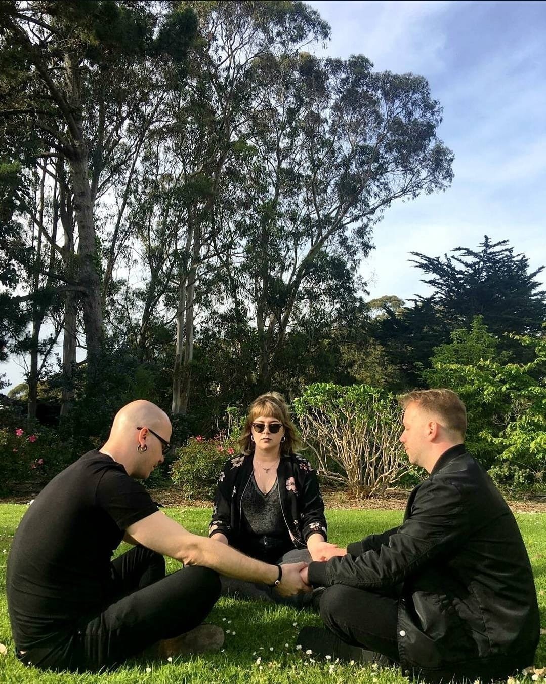 Shea Bilé and two others sit on grass in a park, holding hands in a circle, with trees and bushes in the background.
