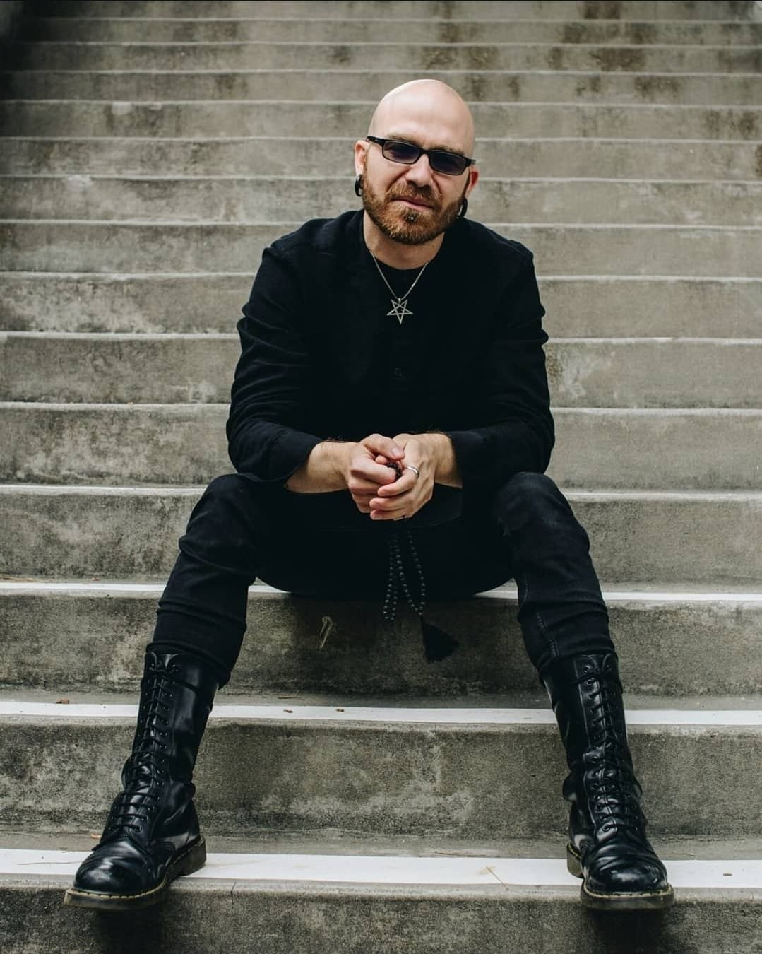Shea Bilé wears an inverted pentagram necklace sitting on concrete stairs, wearing black clothing and boots, with hands clasped. Author portrait style photograph.