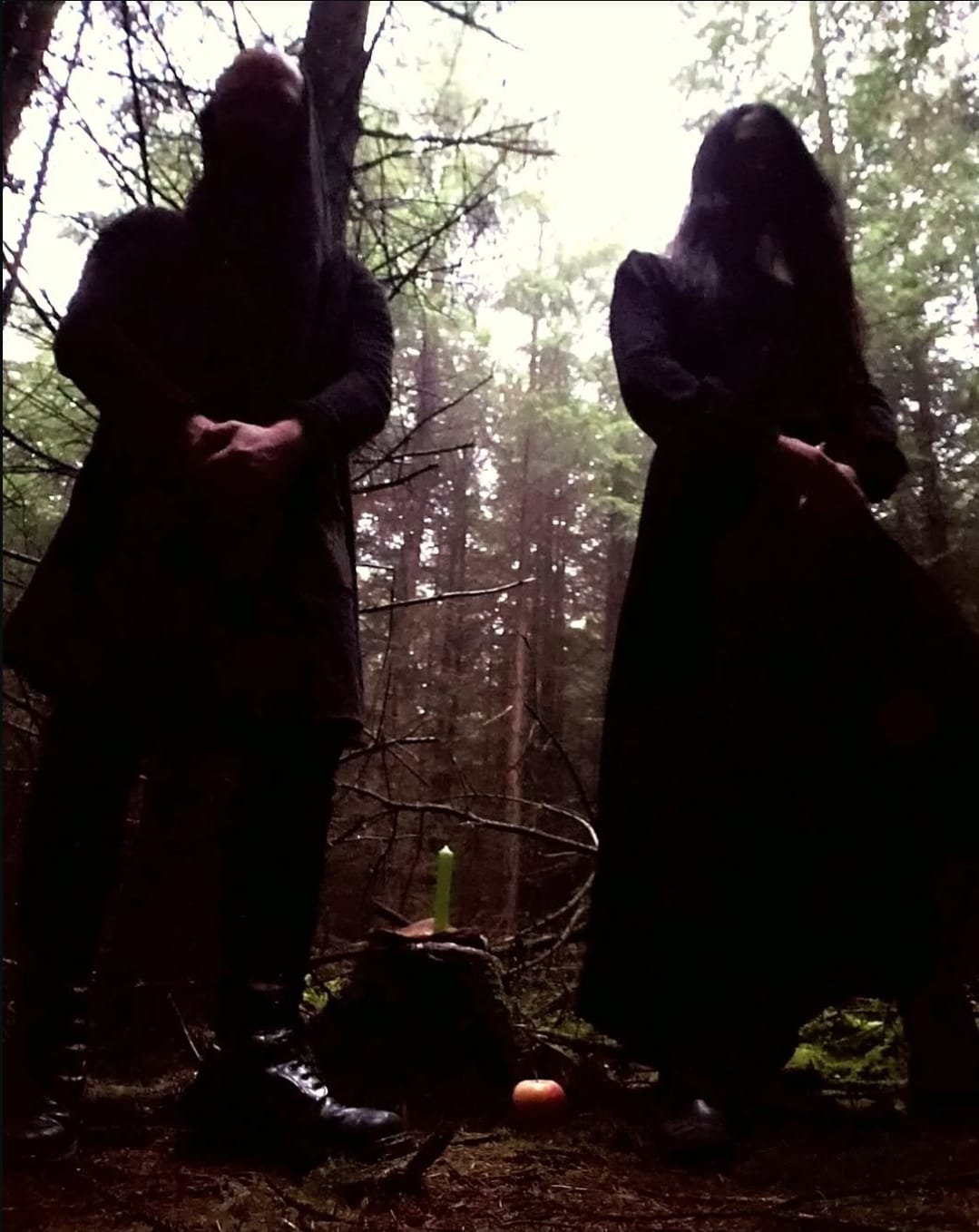 Shea Bilé and Luzia Lowe stand in a dark forest, both dressed in black, facing the camera with hands clasped in front. There are two candles on the ground, one lit, along with an apple placed nearby. Wicklow, Ireland.