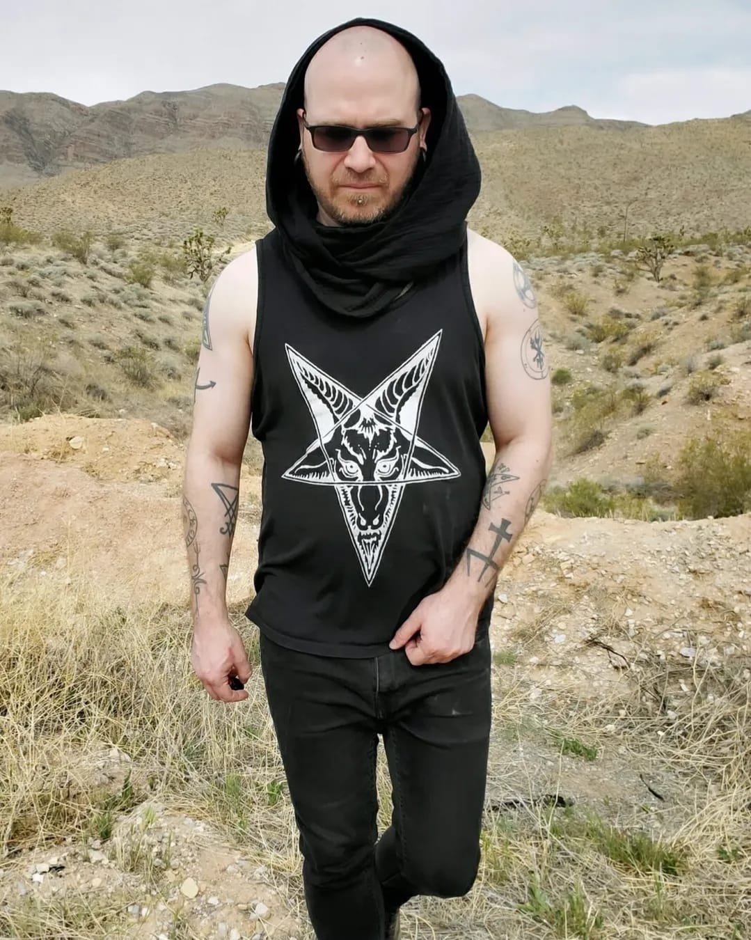 Shea Bilé is wearing a black tank top with a Baphomet design, standing in a desert landscape with mountains in the background.