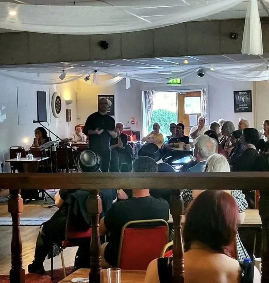 Shea Bilé is standing and speaking to an audience in a dimly lit room with tables and chairs, Pandaemonium UK, Weymouth, England. 