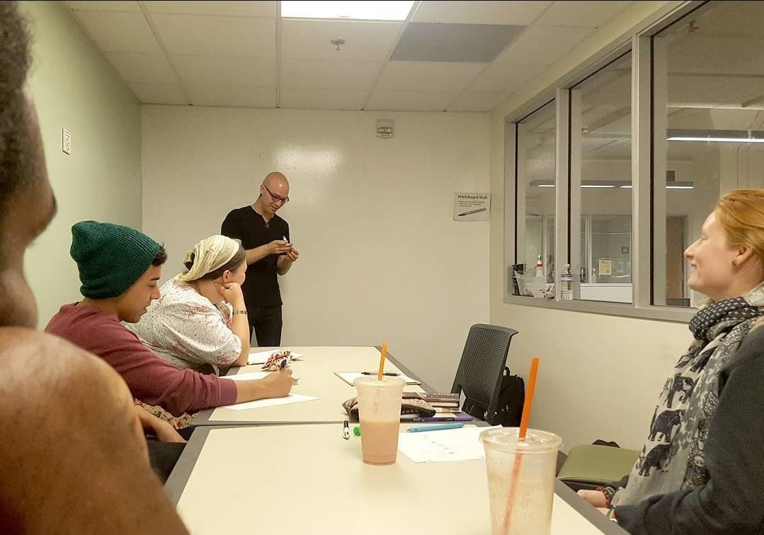 Group of people in a meeting room, sitting at a table with drinks and notebooks, while two people are near the wall, one standing and the other seated and smiling. Shea Bilé gives a lecture in front of a large white board. Occult Student Alliance.
