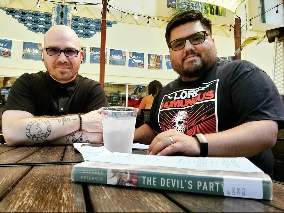 Shea Bilé and Dr. William Chavez sitting at a wooden table outdoors, with drinks, a book titled 'The Devil's Party.' Background has banners with Budweiser and Modelo branding.