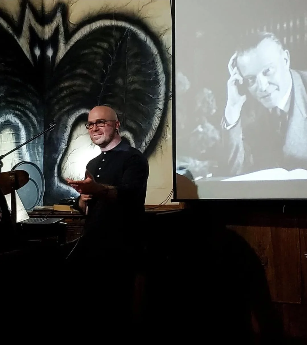 Shea Bilé stands in front of a microphone, gesturing with his hand, in a room with a large mural of a bat, a projector slide shows an image of Hanns Hienz Ewers. The Fox Tavern. New York City.