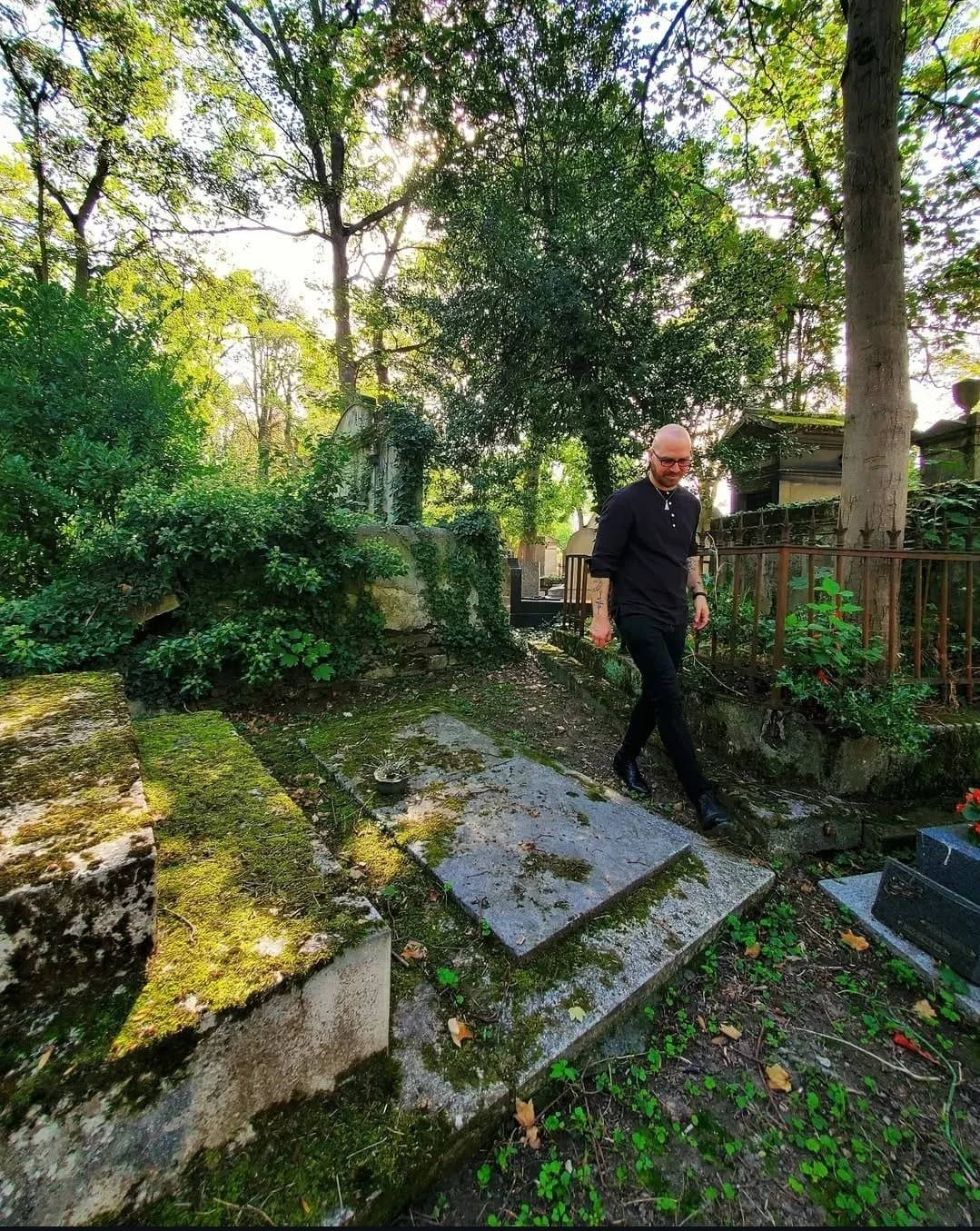 Shea Bilé walking through a wooded graveyard with moss-covered tombstones and trees in the background. Paris, France.