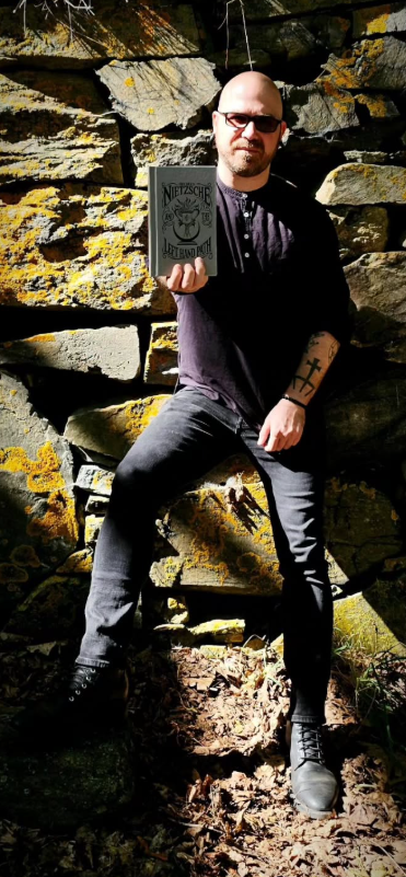 Author Shea Bilé (Shea Bile) seated outdoors against a stone wall, holding a copy of Friedrich Nietzsche and the Left Hand Path in natural light.