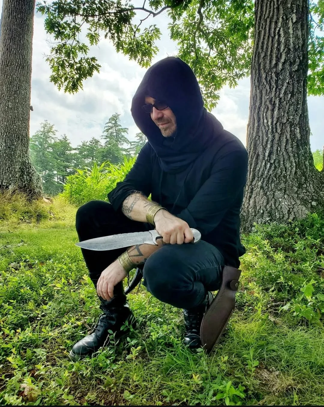 Shea Bilé wears a black hooded cloak, and combat boots, is sitting outdoors surrounded by trees and greenery, holding a knife. Renaissance Faire in Connecticut. 