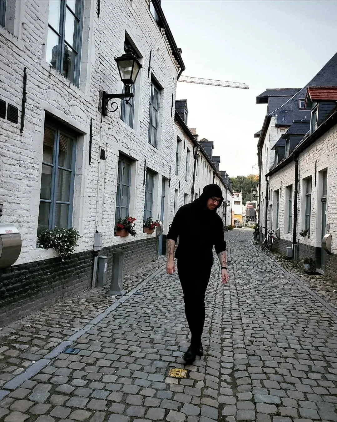 Shea Bilé dressed in black walking on cobblestone street between white brick buildings with flowerpots, bicycles, and street lamps. Leuven, Belgium.
