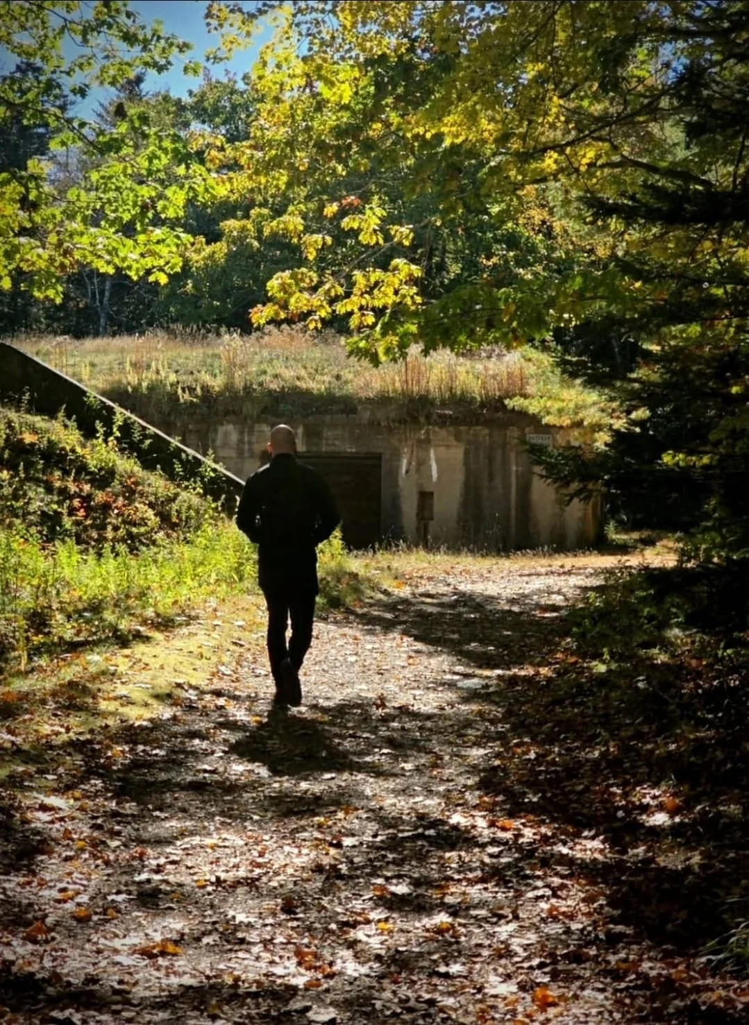Shea Bilé dressed in black walking down a dirt trail surrounded by trees with yellow and green leaves, sunlight filtering through the branches. Approaching an abandoned bunker. Maine.