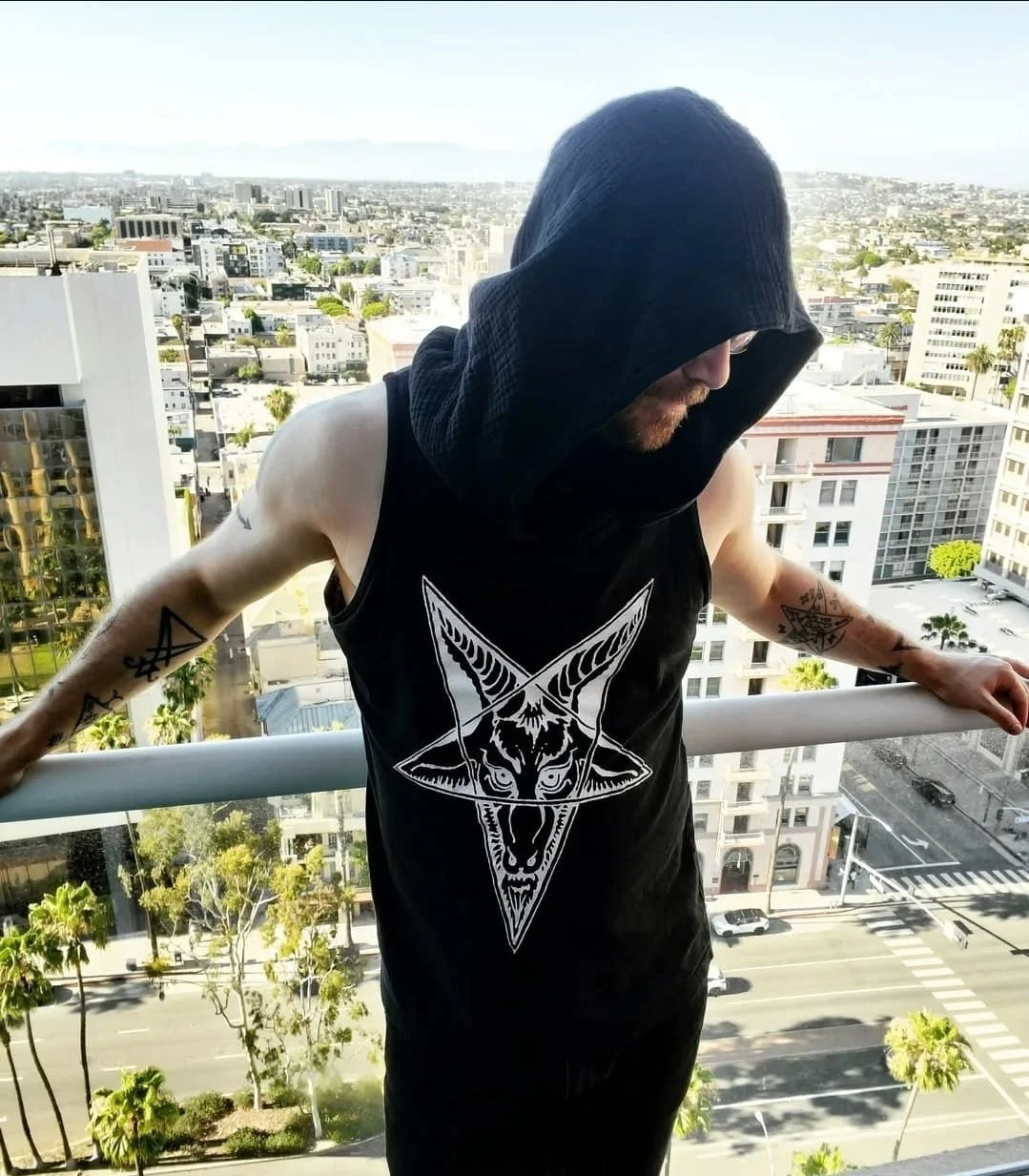 Shea Bilé is wearing a black sleeveless shirt with a Baphomet design, on a balcony overlooking the Los Angeles cityscape with tall buildings and trees, his face partially hidden by a large black hood or cloth.